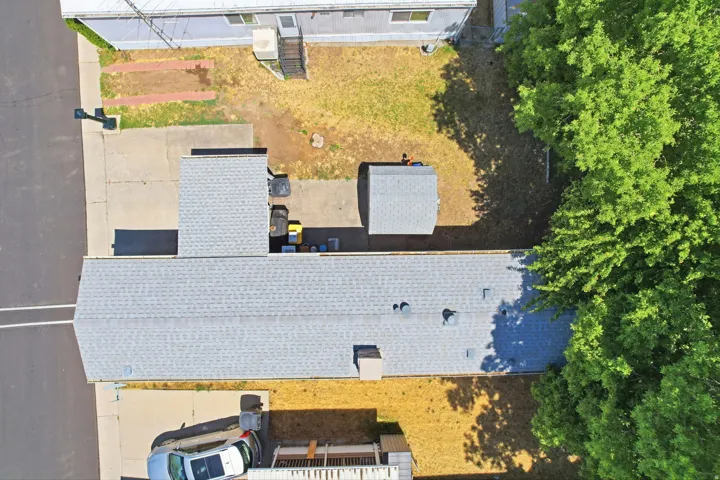 Aerial view of the property featuring a light grey shingle roof, a detached shed with a light grey shingle roof, paved driveway, concrete patio, and mature trees