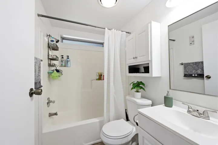 Bright bathroom featuring a white tub and shower combination with an integrated window, a white vanity with a single basin sink, and a wall-mounted storage cabinet