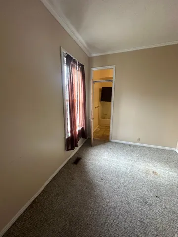 Carpeted room featuring a window with sheer curtains, crown molding, and an interior door revealing a bathroom with a shower/tub combination
