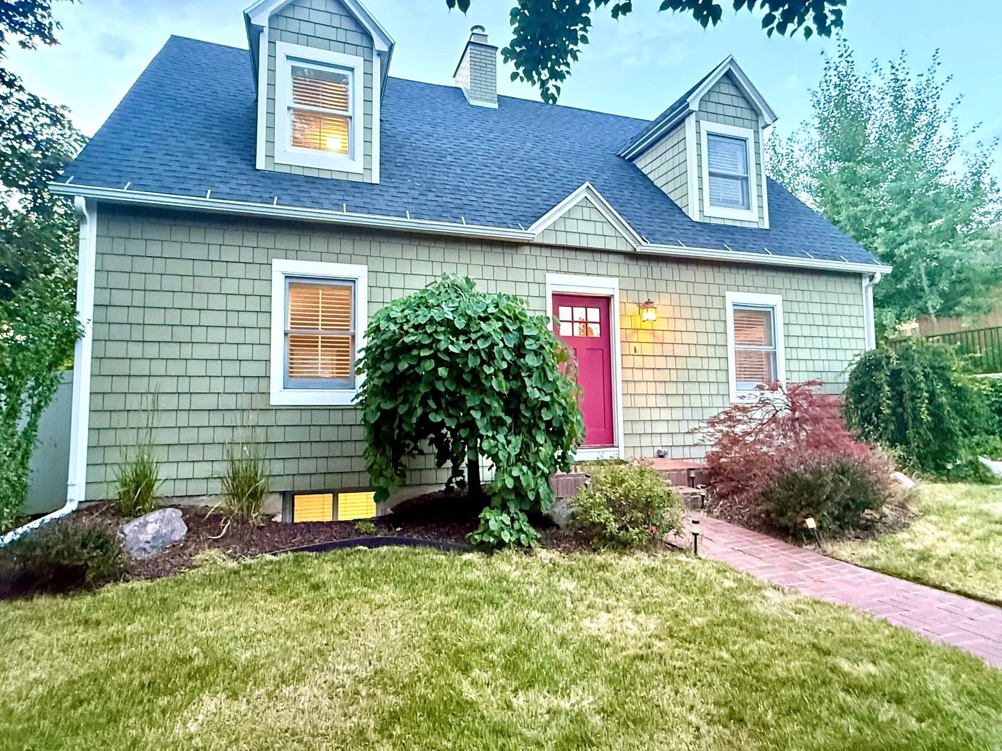 Dormered dwelling with green shingle siding, a red front door, and a brick pathway