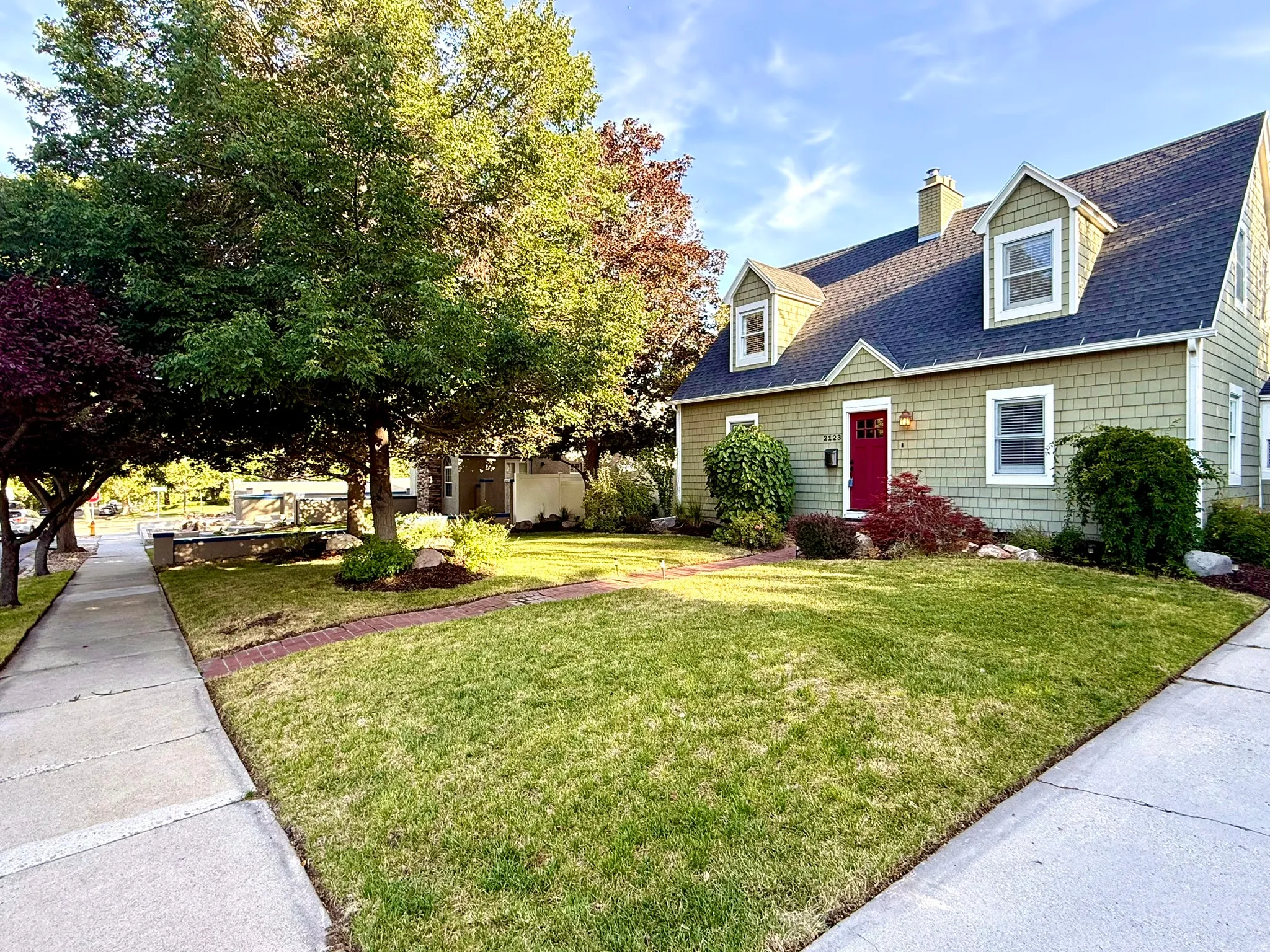 Gable roofline residence featuring light green shake siding, a red entry door, multiple dormer windows, and a brick-lined walkway