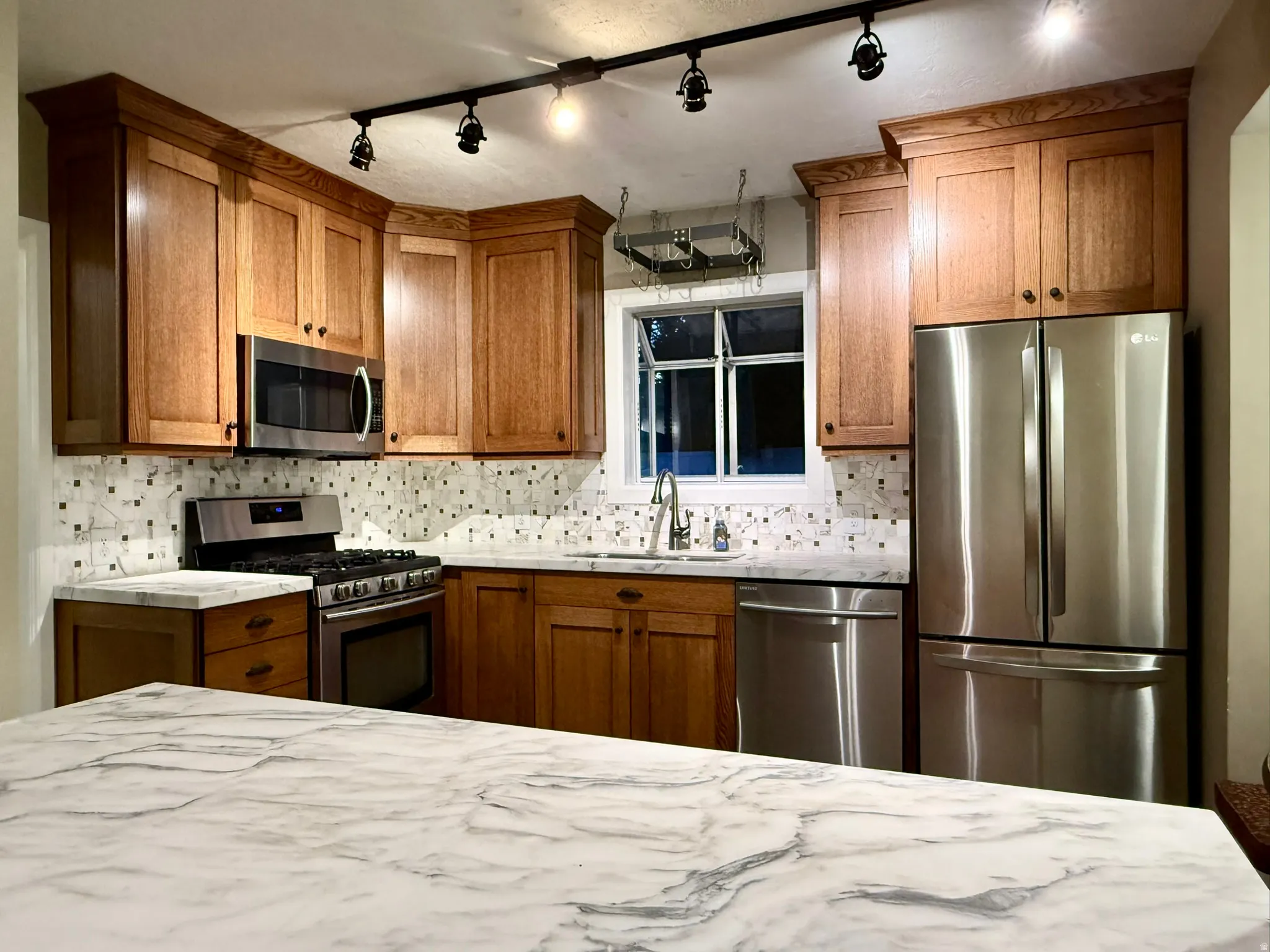 Modern kitchen featuring a white and grey veined countertop, stainless steel appliances, wood cabinetry, track lighting, and a patterned tile backsplash