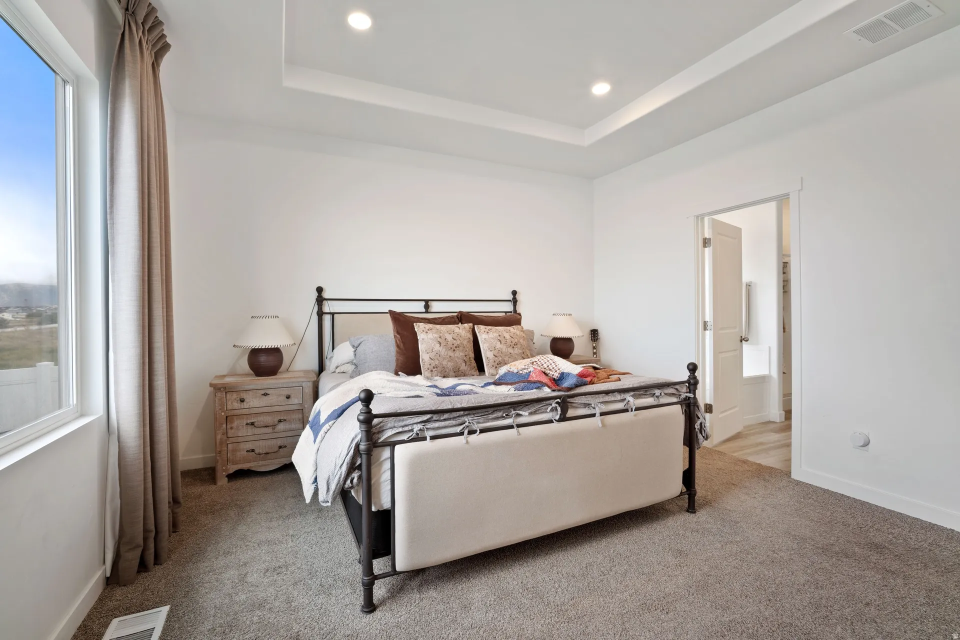 Spacious bedroom featuring a recessed ceiling with integrated lighting, a large window, light-colored carpeting, and a doorway opening to an additional room