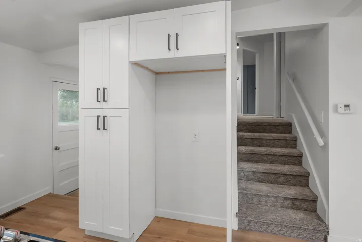 White shaker-style cabinetry with matte black hardware, integrated refrigerator alcove, wood-finish flooring, and carpeted staircase with white handrail