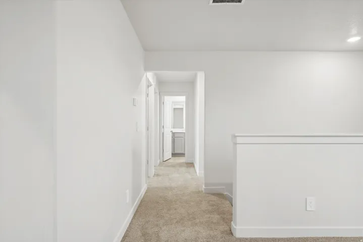 **DISCLAIMER** Interior package may vary. Carpeted hallway with white walls and trim, leading to a bathroom with a vanity and mirror