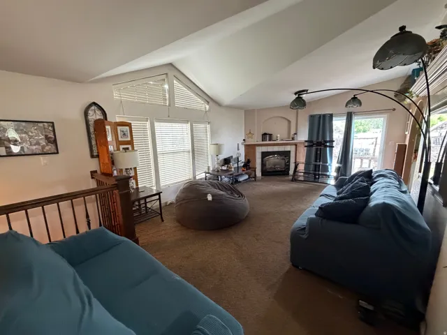 Spacious living area featuring a vaulted ceiling, multiple windows with blinds, a gas fireplace with a white tile surround, and a light brown carpet