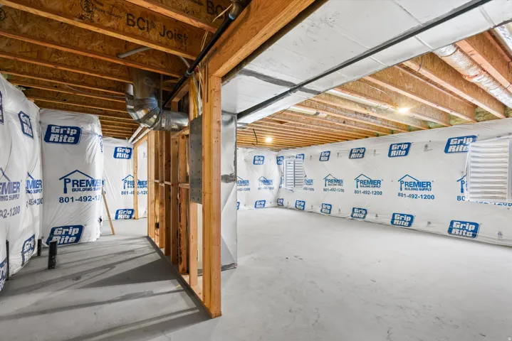 Unfinished basement featuring exposed wood joists and subflooring, concrete flooring, and perimeter walls framed with wood studs and covered in building wrap