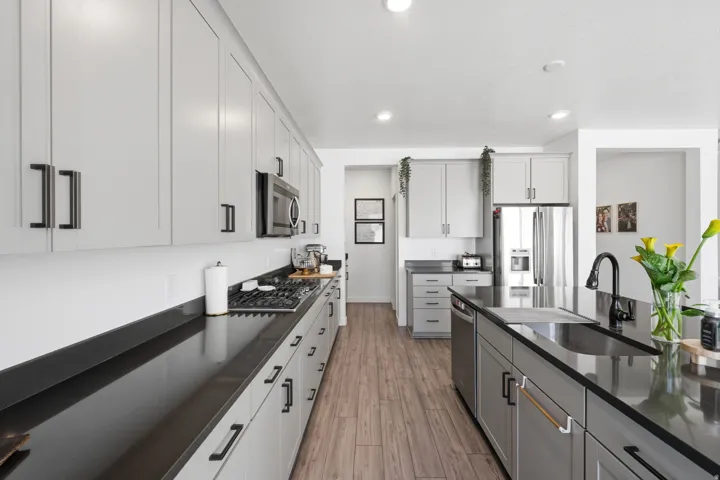 Contemporary kitchen featuring light gray cabinetry with black hardware, dark countertops, wood-finish flooring, a stainless steel refrigerator, and a kitchen island with an integrated sink and black faucet