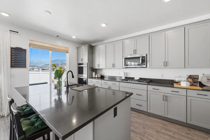 Kitchen featuring a large central island with an undermount sink, light gray cabinetry, dark countertops, built-in stainless steel appliances, and wood-finish flooring