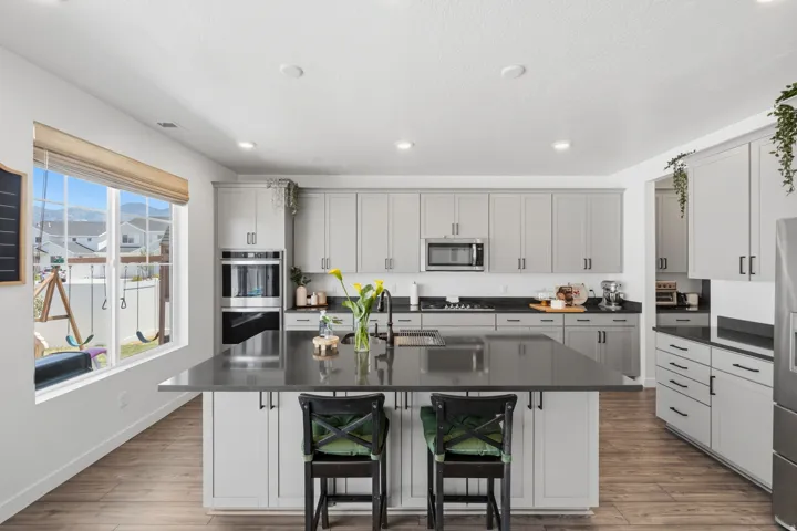 Spacious kitchen featuring an expansive center island with seating, a suite of stainless steel appliances including a built-in oven and microwave, abundant light gray cabinetry, dark countertops, and wood-finish flooring