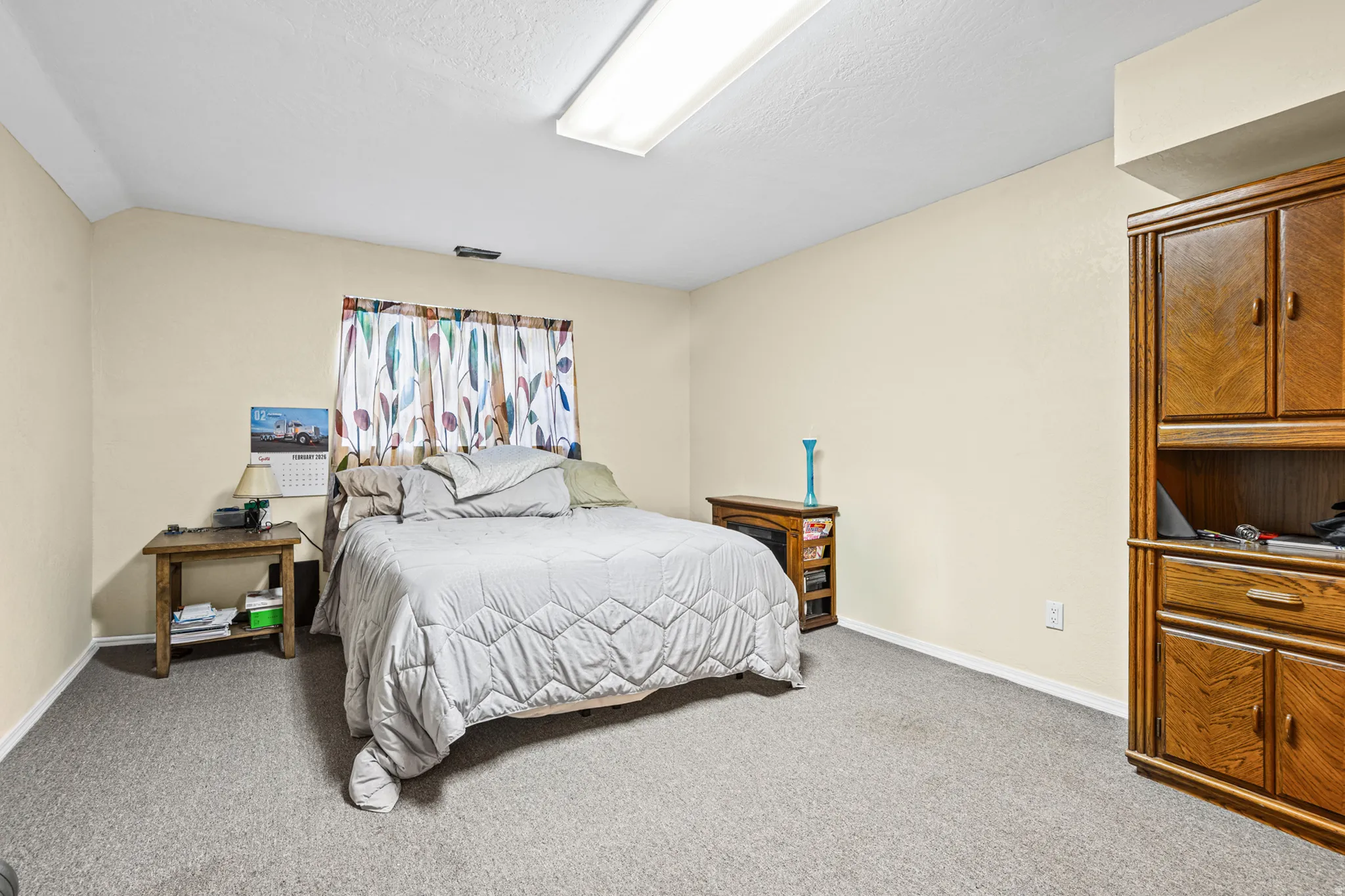 Carpeted room featuring a window with patterned curtains, neutral wall paint, and a built-in wood-finish cabinet with shelving and drawers