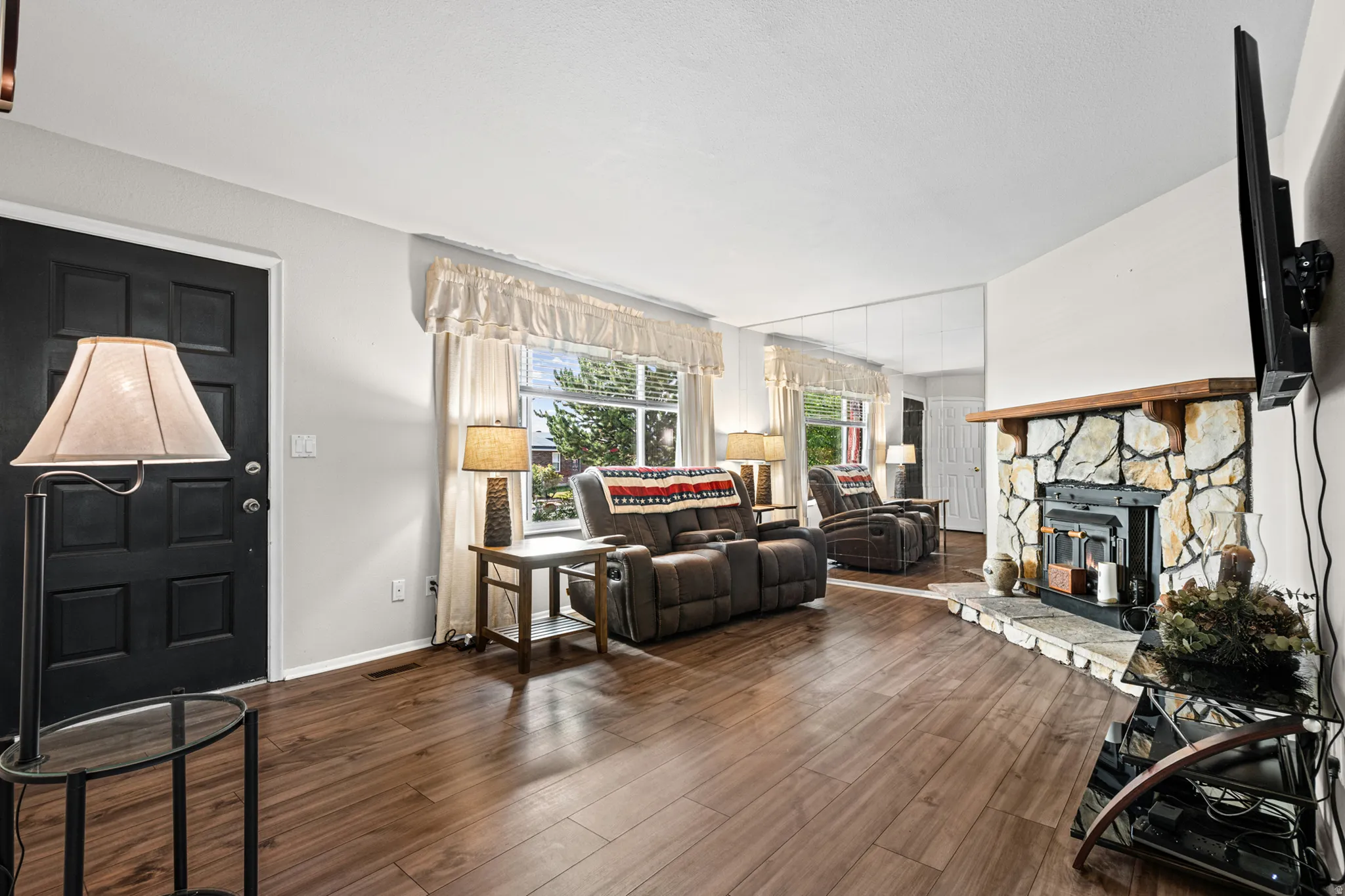 Living area featuring wood-finish flooring, a stone-clad fireplace with a wood mantle, and multiple windows with drapery