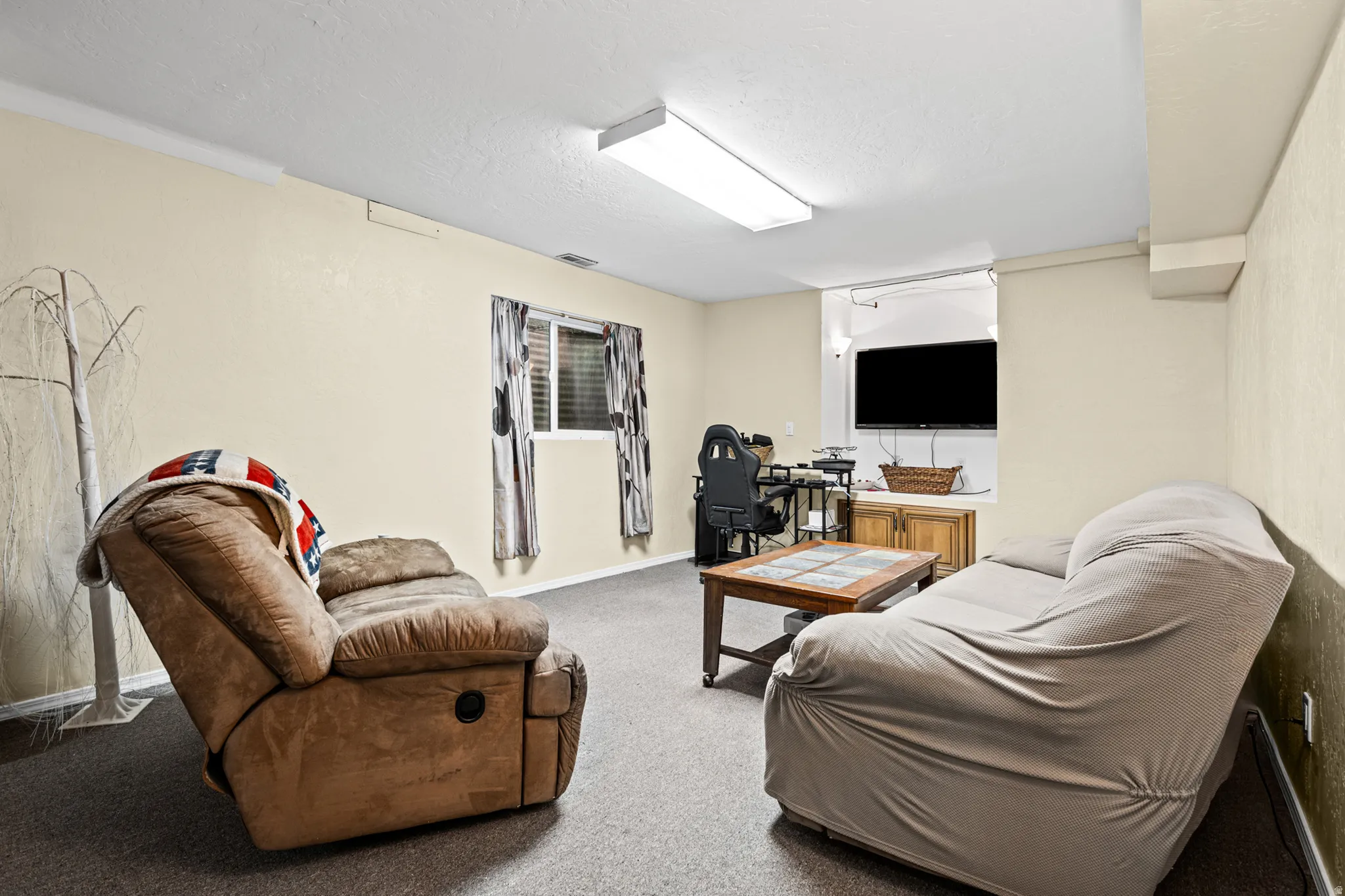 Recessed entertainment area with built-in cabinetry, neutral wall color, window with horizontal blinds, overhead fluorescent lighting, and carpeted flooring