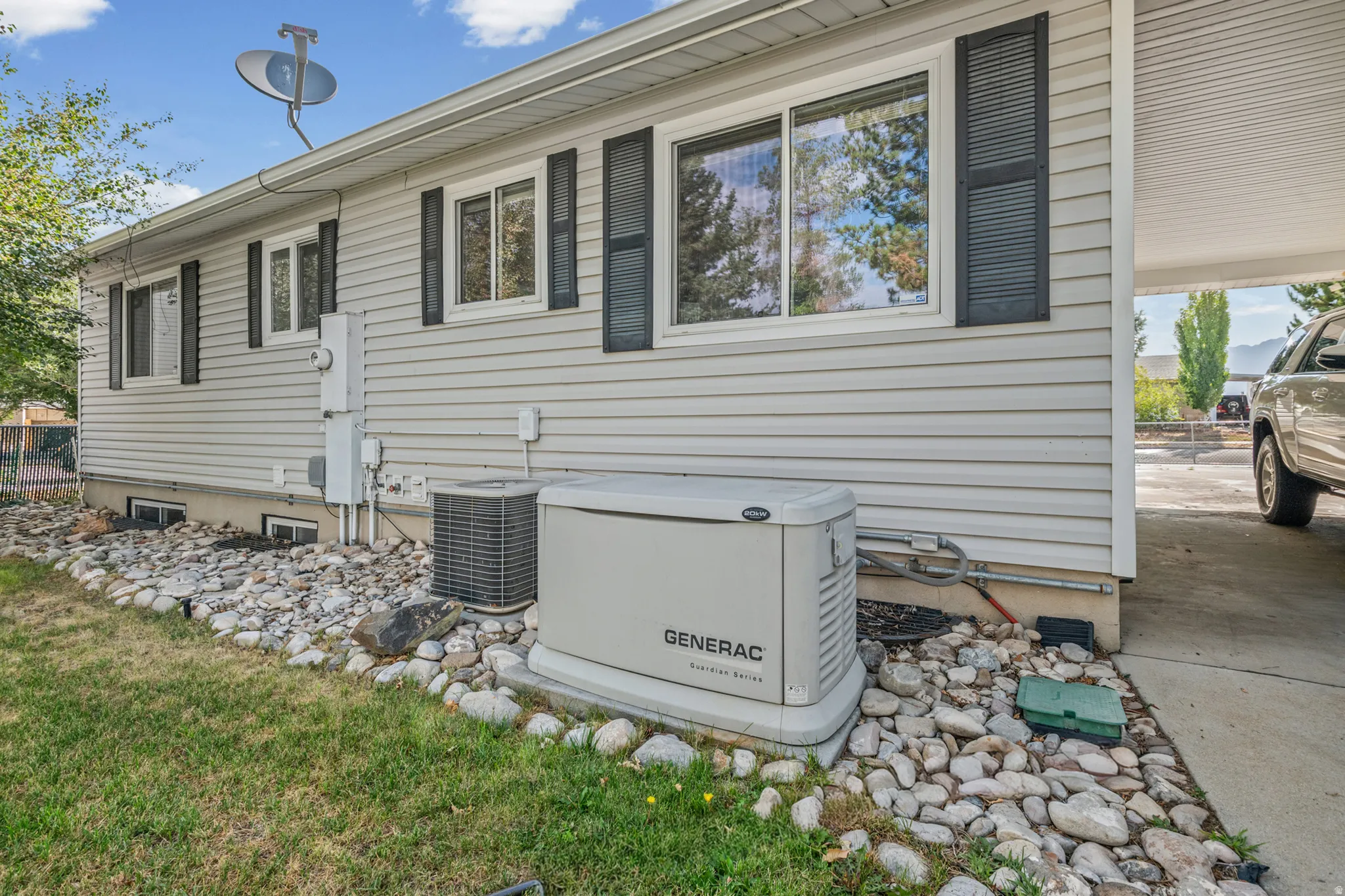 Exterior siding with multiple windows featuring dark louvered shutters, a gravel landscaping bed, and a Generac whole-house generator