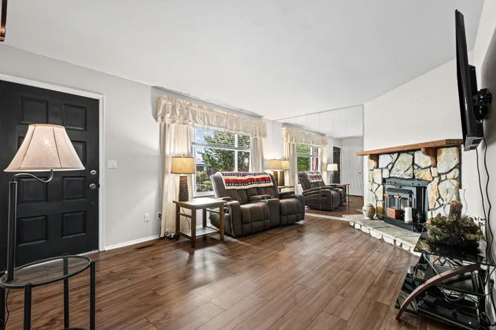 Living area featuring wood-finish flooring, a stone-clad fireplace with a wood mantle, and multiple windows with drapery