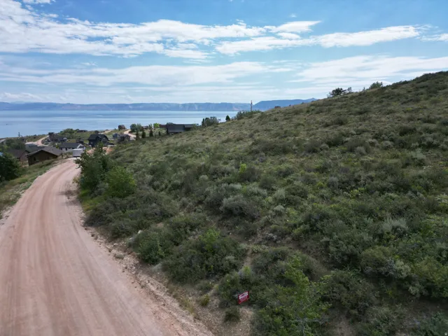 Expansive sloped terrain with natural shrubbery, featuring a visible dirt road, distant water body, and scattered residential structures