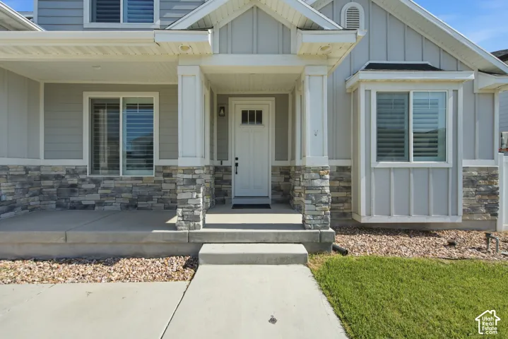 Light gray siding exterior with stone veneer accents, a covered concrete porch, white paneled front door with a transom window, and a bay window featuring louvered blinds