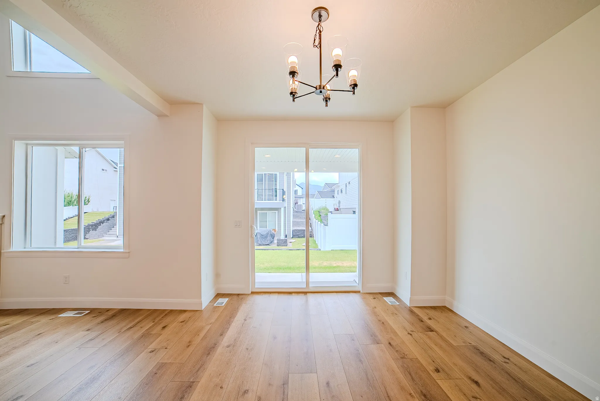 Spacious room featuring wood-finish flooring, a contemporary chandelier, white walls, and a large sliding glass door opening to a manicured lawn