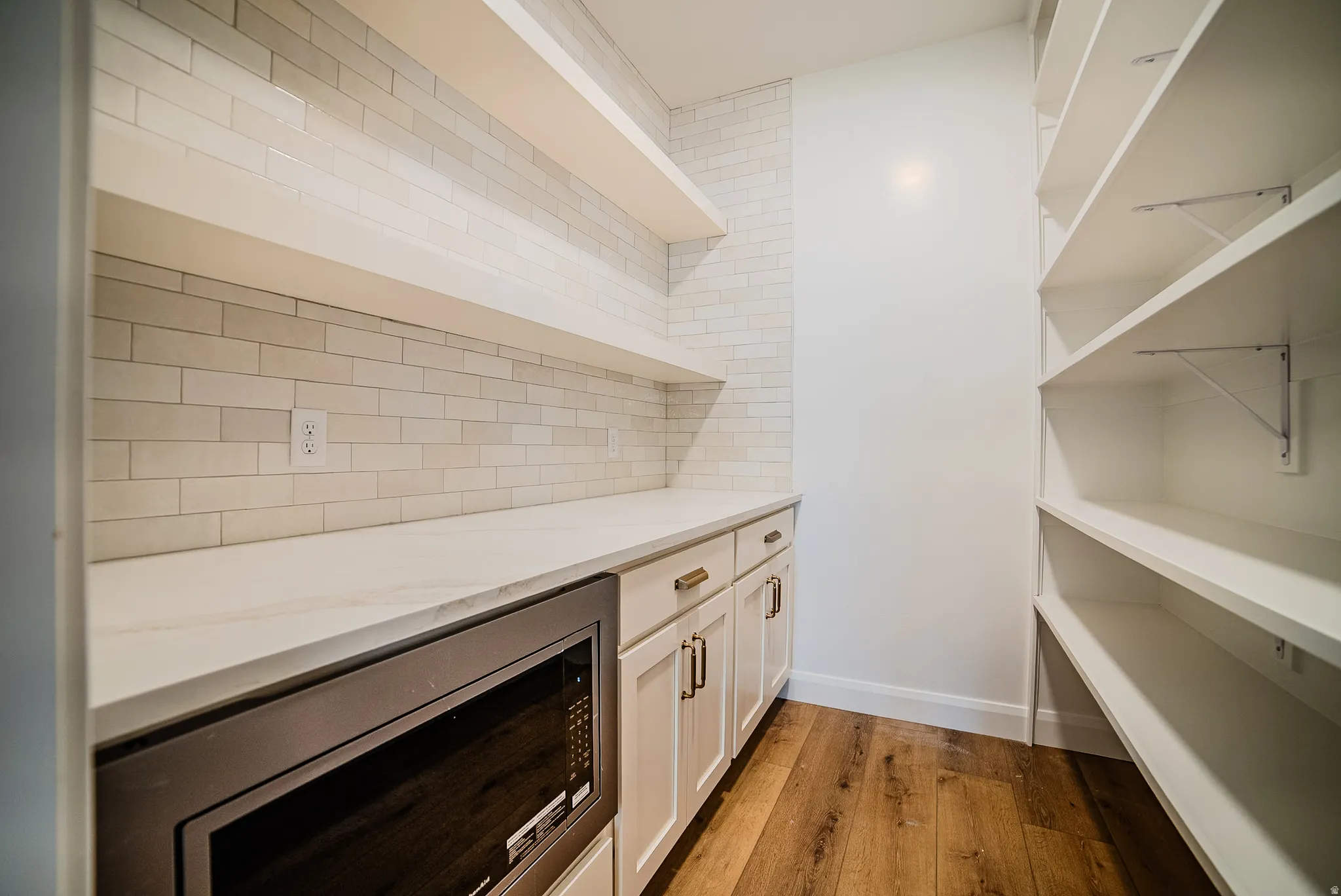 Kitchen pantry featuring a built-in microwave, white shaker cabinetry with brass hardware, a solid surface countertop, white subway tile backsplash, open shelving, and wood-finish flooring