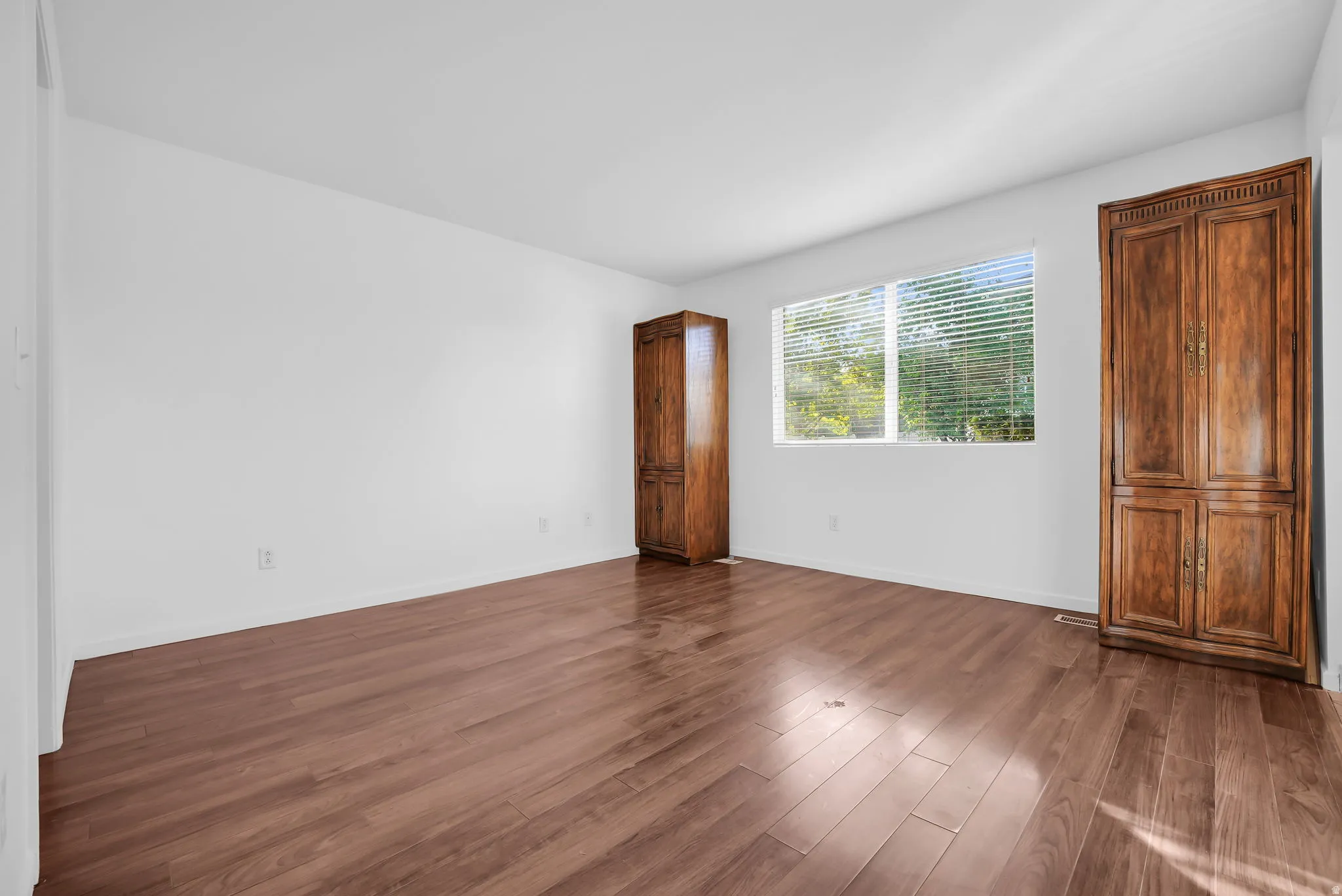 Spacious room featuring wood-finish flooring, white walls, and a large window with blinds