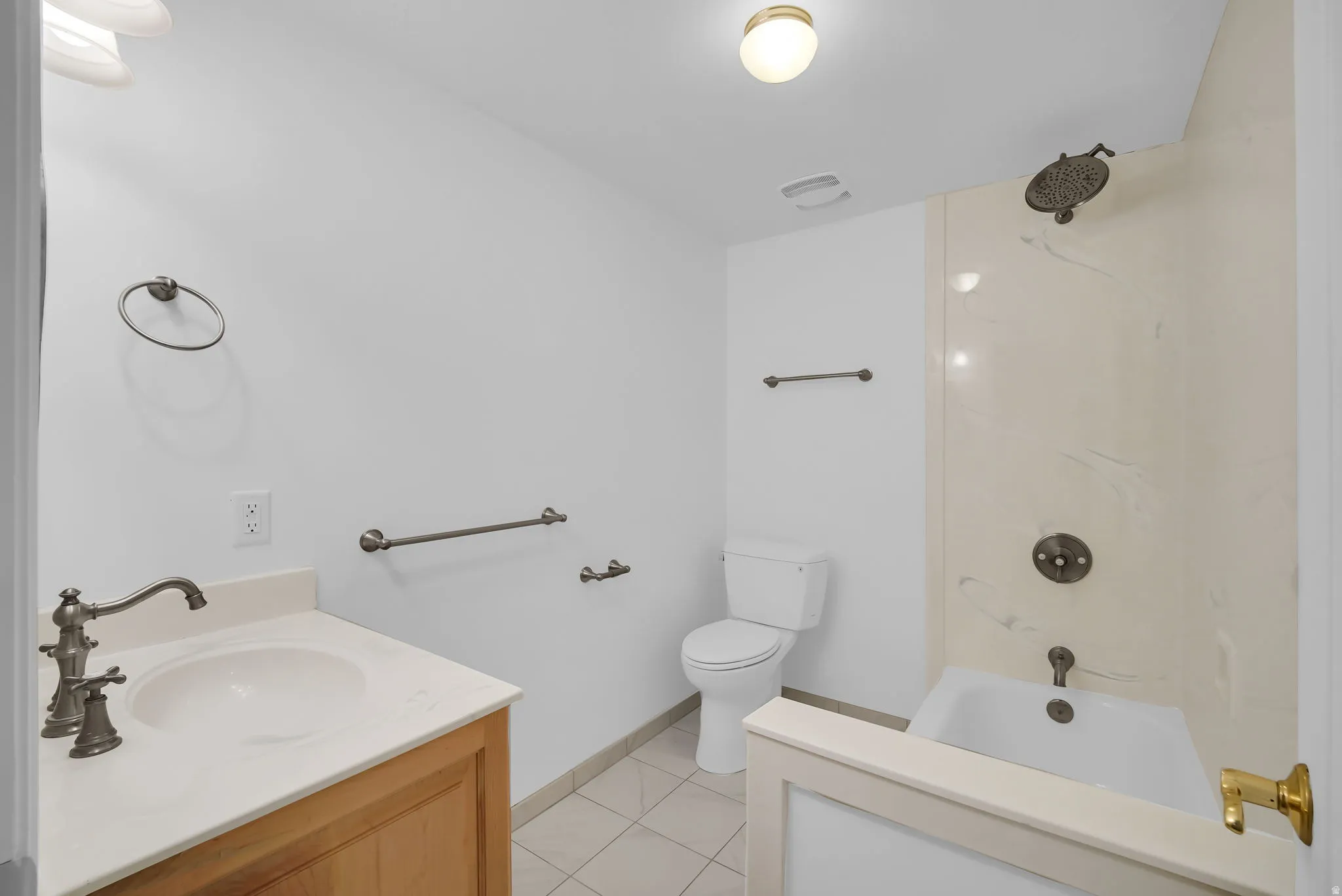 Bathroom featuring a wood vanity with a single basin sink and bronze-finish faucet, a toilet, a shower-over-tub combination with a wall-mounted showerhead, and a tiled floor