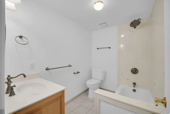 Bathroom featuring a wood vanity with a single basin sink and bronze-finish faucet, a toilet, a shower-over-tub combination with a wall-mounted showerhead, and a tiled floor