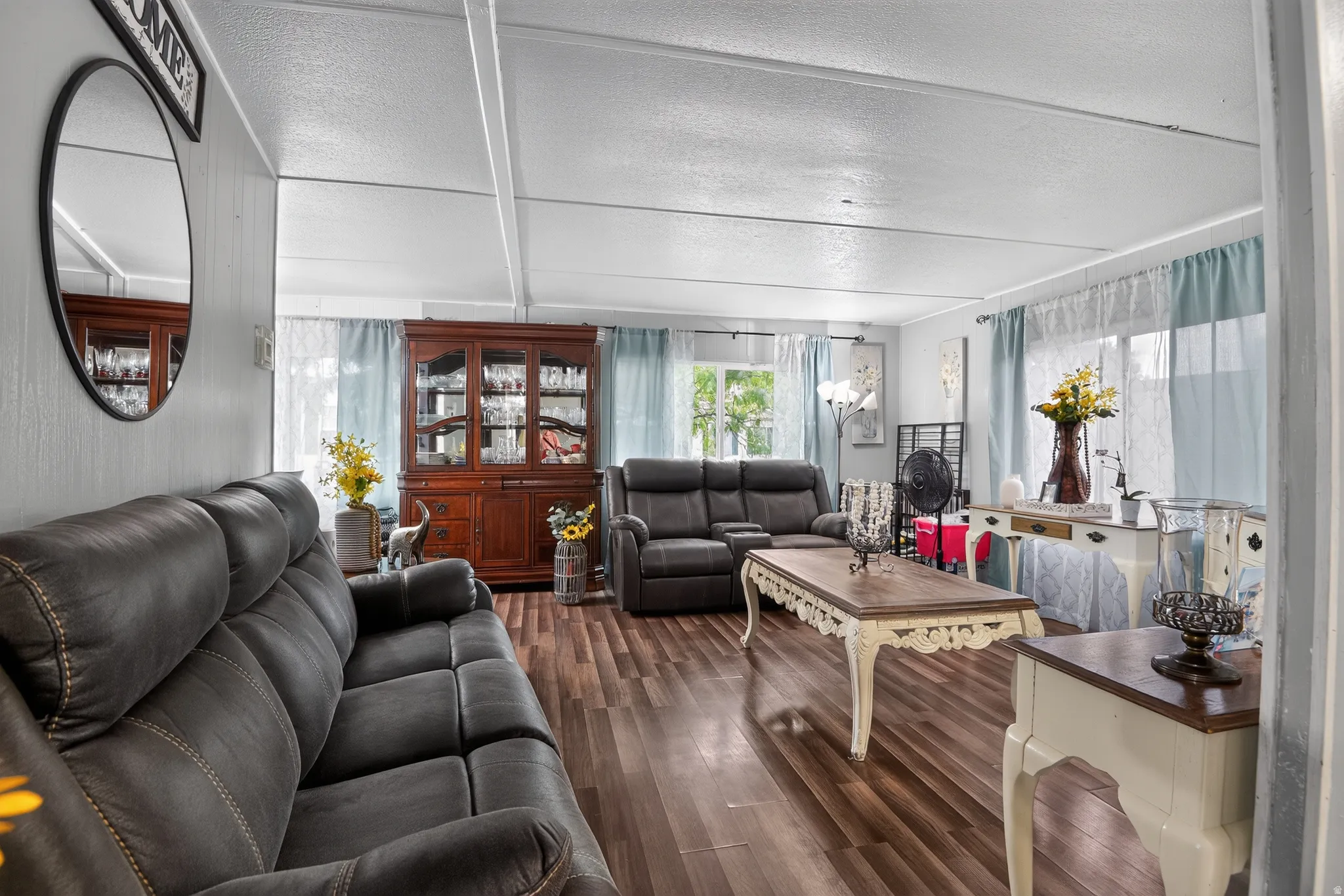 Spacious living area featuring wood-finish flooring, light-colored walls, and multiple windows with light-filtering curtains