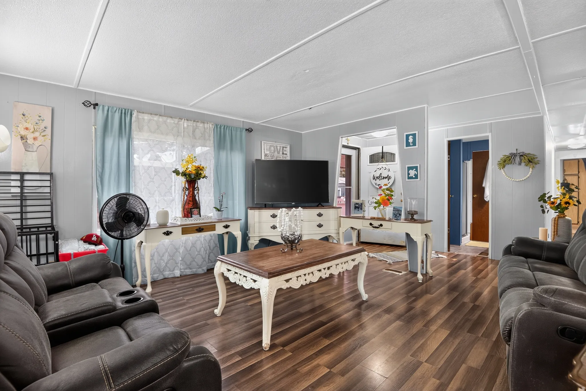 Spacious living area featuring wood-finish flooring, light gray wall paneling, and a bright window with light blue drapery