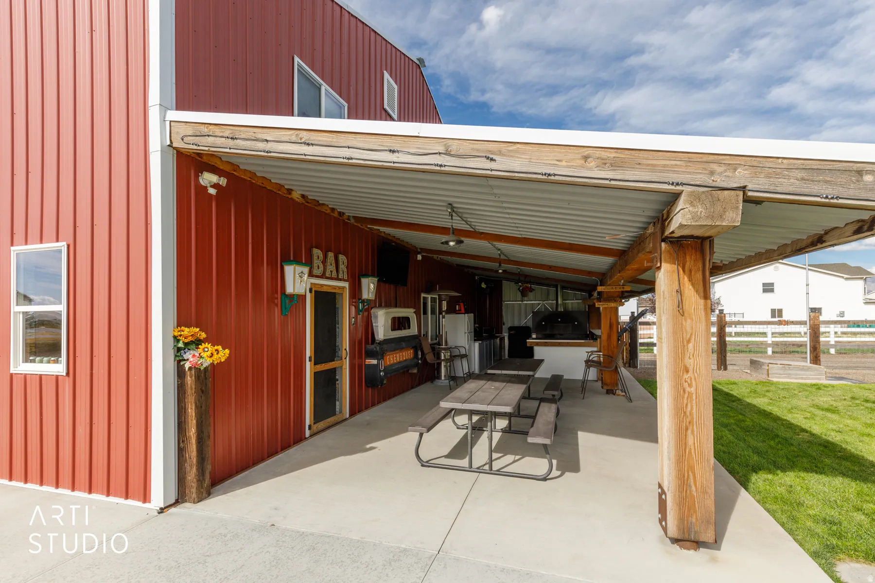 Expansive covered patio featuring a full outdoor kitchen, wood beam supports, concrete flooring, and corrugated metal roofing