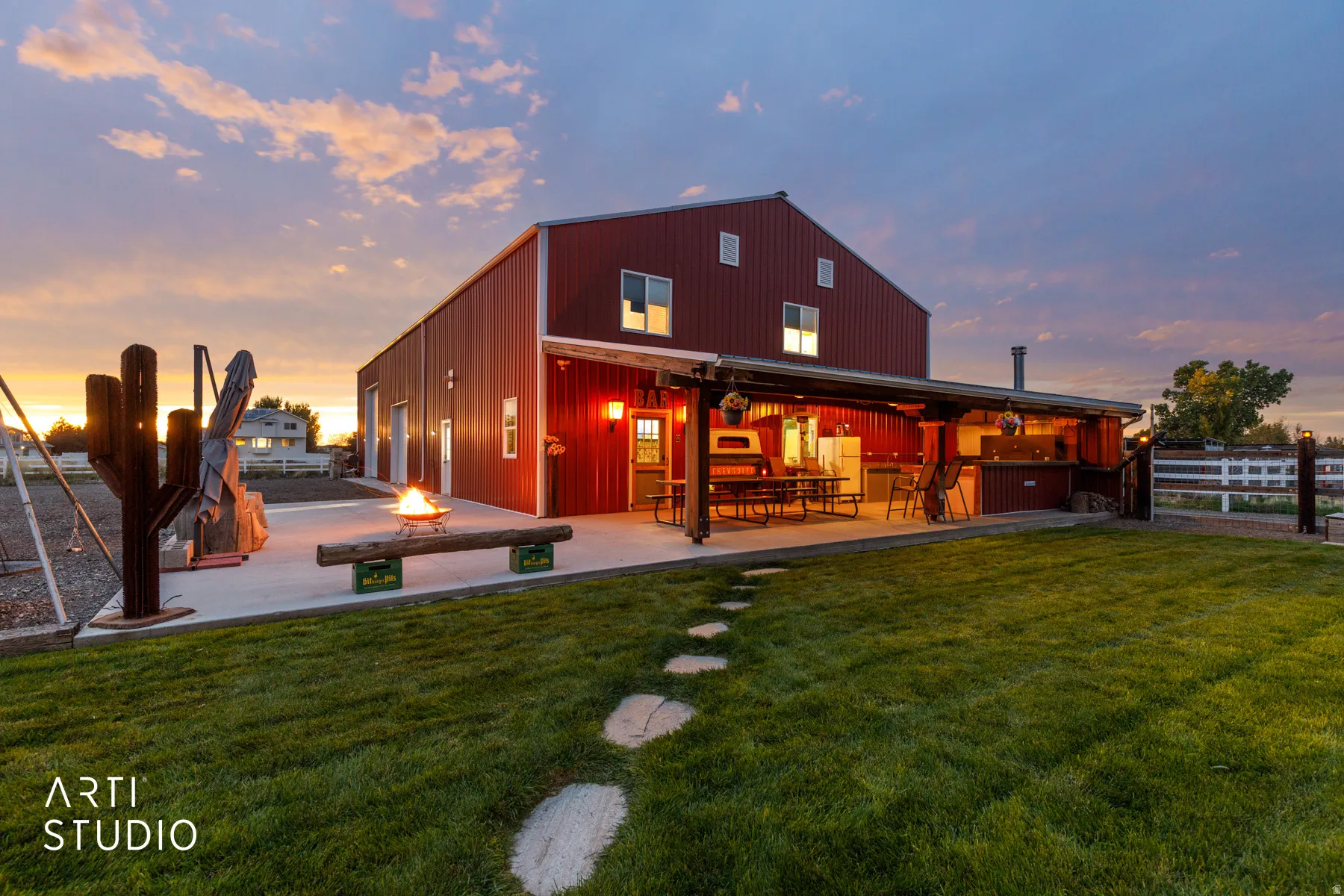 Red metal barn-style structure featuring a covered patio with an outdoor kitchen, a stone pathway, a lush green lawn, and a fire pit area