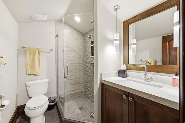 Contemporary bathroom featuring a glass-enclosed shower with tile surround and floor, a vanity with a light-toned countertop and integrated sink, a wood-framed mirror, and modern pendant lighting