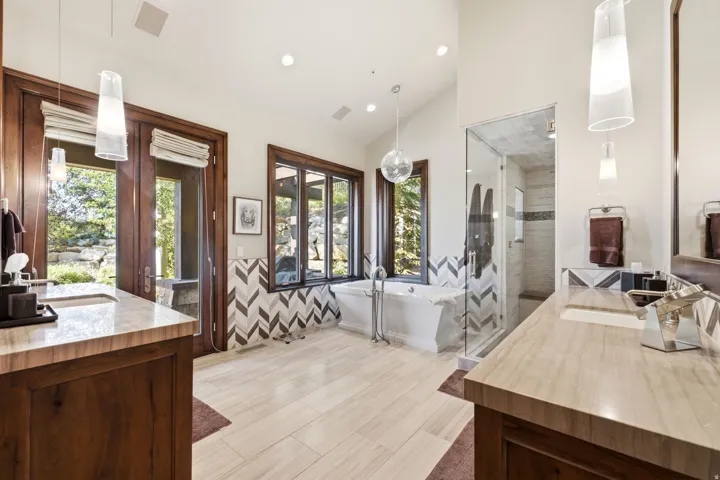 Spacious bathroom featuring a freestanding tub, glass-enclosed shower, two wood vanities with integrated sinks, light-toned flooring, and chevron-patterned wall tiling