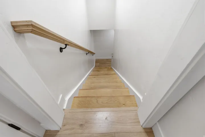 Staircase featuring light wood-finish treads, a contrasting natural wood handrail, matte black handrail supports, and white painted walls