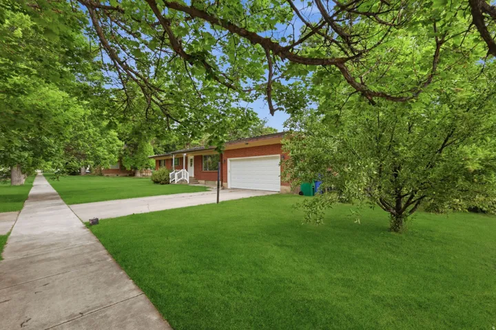 Brick exterior featuring a covered front porch with white railing, a white garage door, a paved driveway, a concrete sidewalk, and mature landscaping
