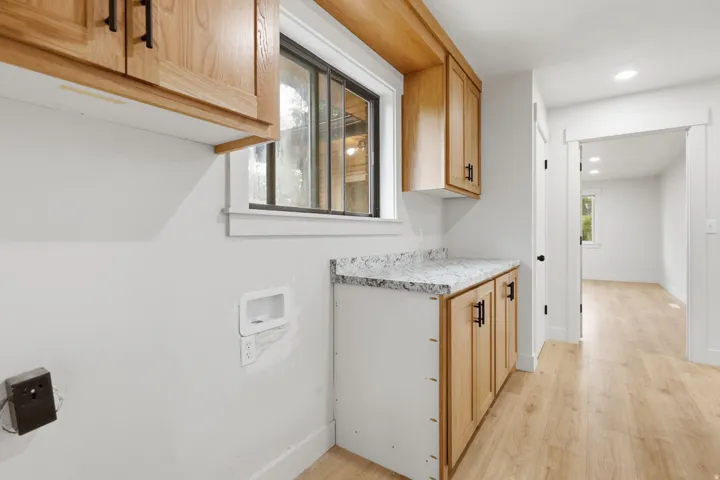 Laundry area featuring light wood-finish cabinetry with matte black hardware, a light-toned speckled countertop, and a window providing natural light