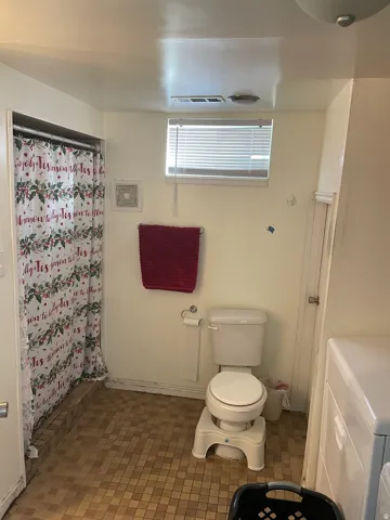 Bathroom featuring a shower stall with a tiled surround, a toilet, a window with blinds, and durable tiled flooring
