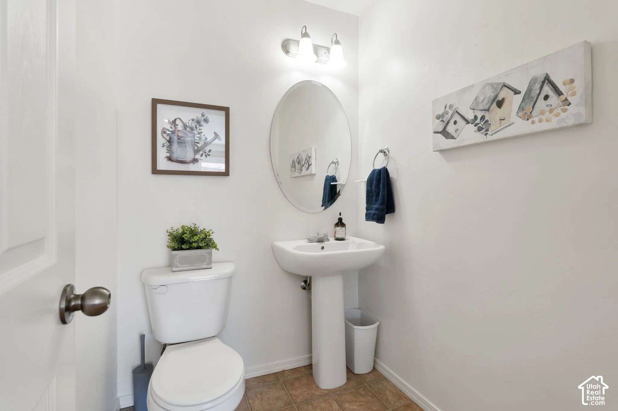 Powder room featuring a pedestal sink, oval mirror, white commode, and warm-toned flooring