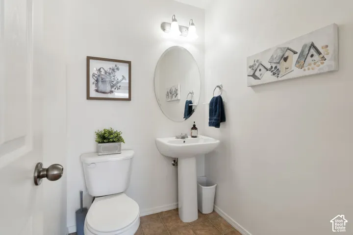 Powder room featuring a pedestal sink, oval mirror, white commode, and warm-toned flooring