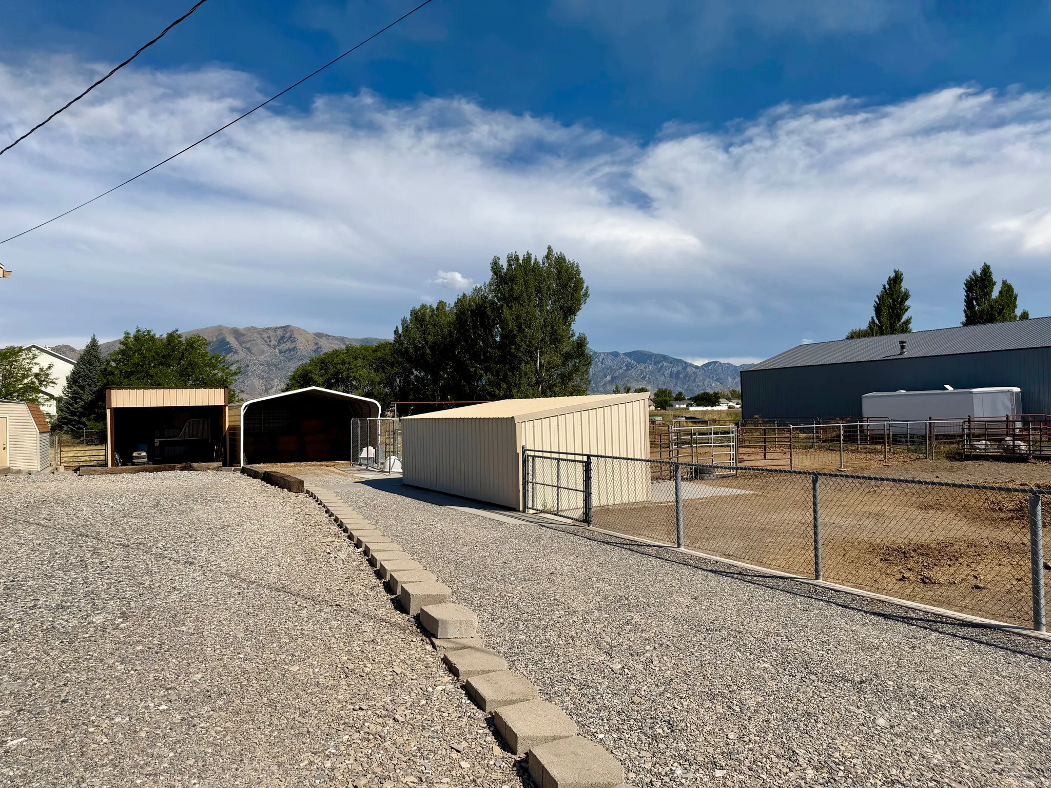 Gravel driveway with concrete paver border, multiple open-sided shelters, and a chain-link fenced area