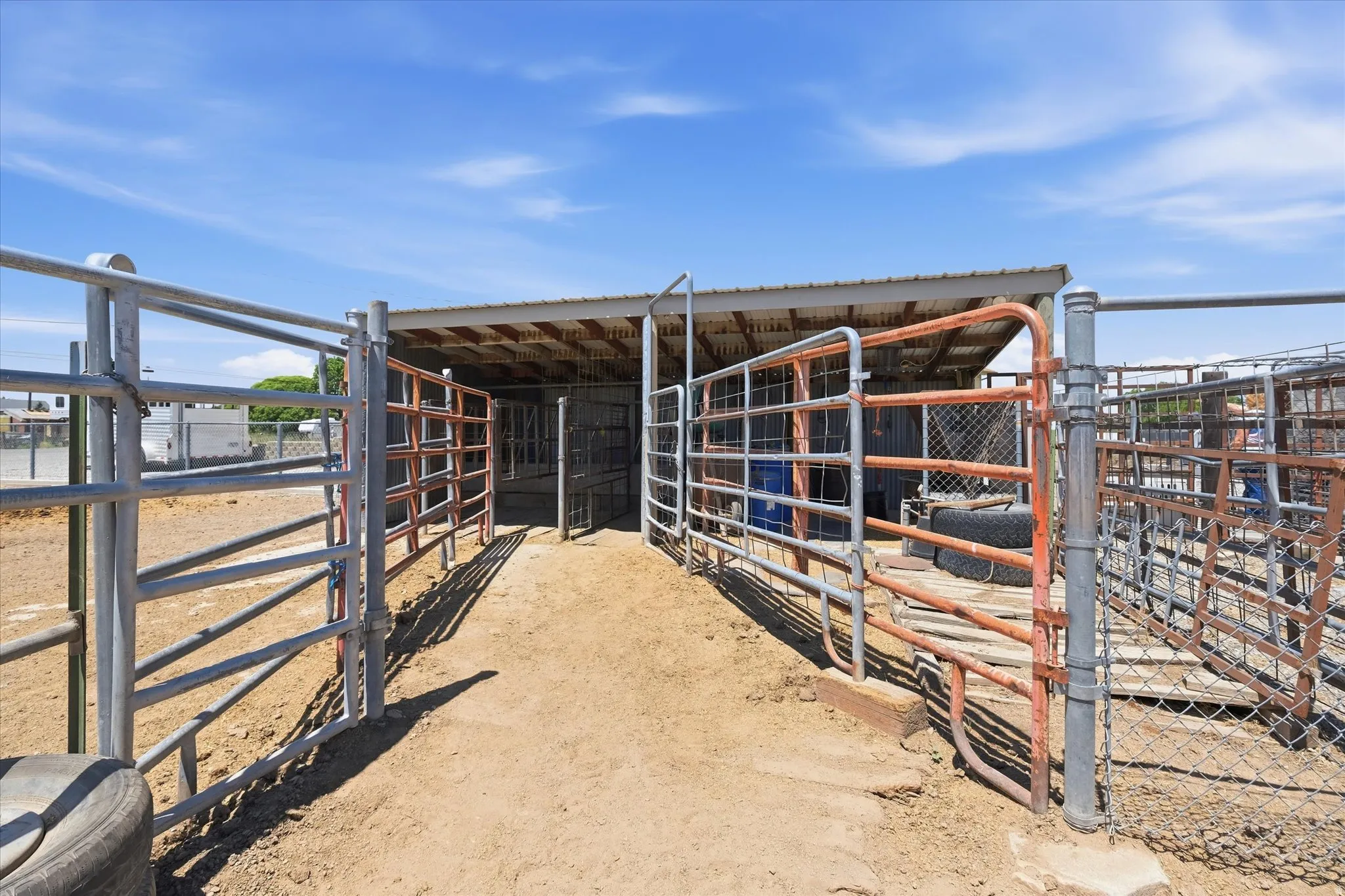 Metal-fenced livestock enclosure featuring multiple gates, a covered shelter, a dirt floor, and chain-link fencing