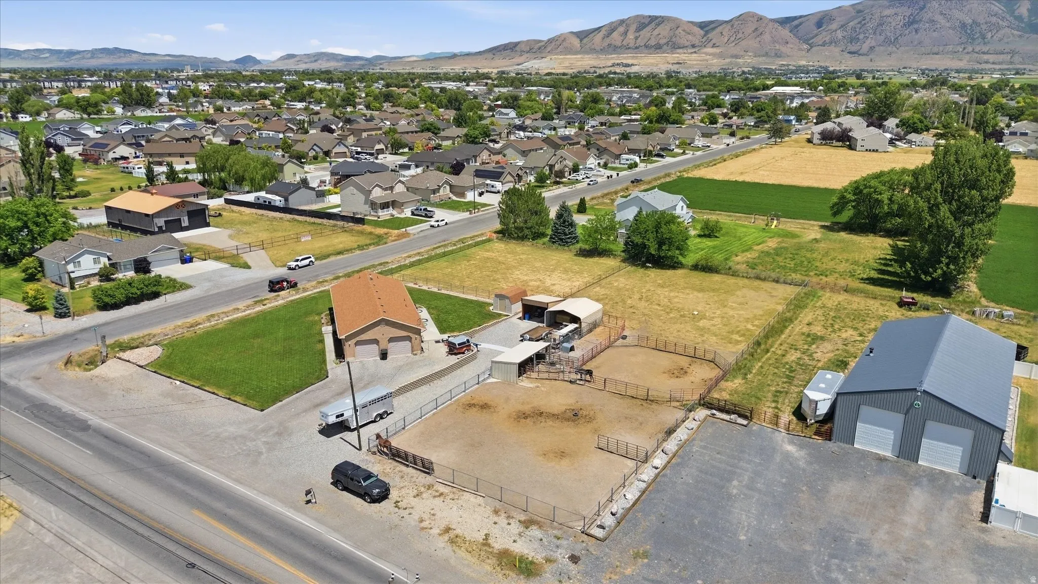 Aerial perspective showcasing a property with a two-story residence featuring an attached garage, a large metal workshop building with dual bay doors, multiple fenced animal enclosures, and substantial land area