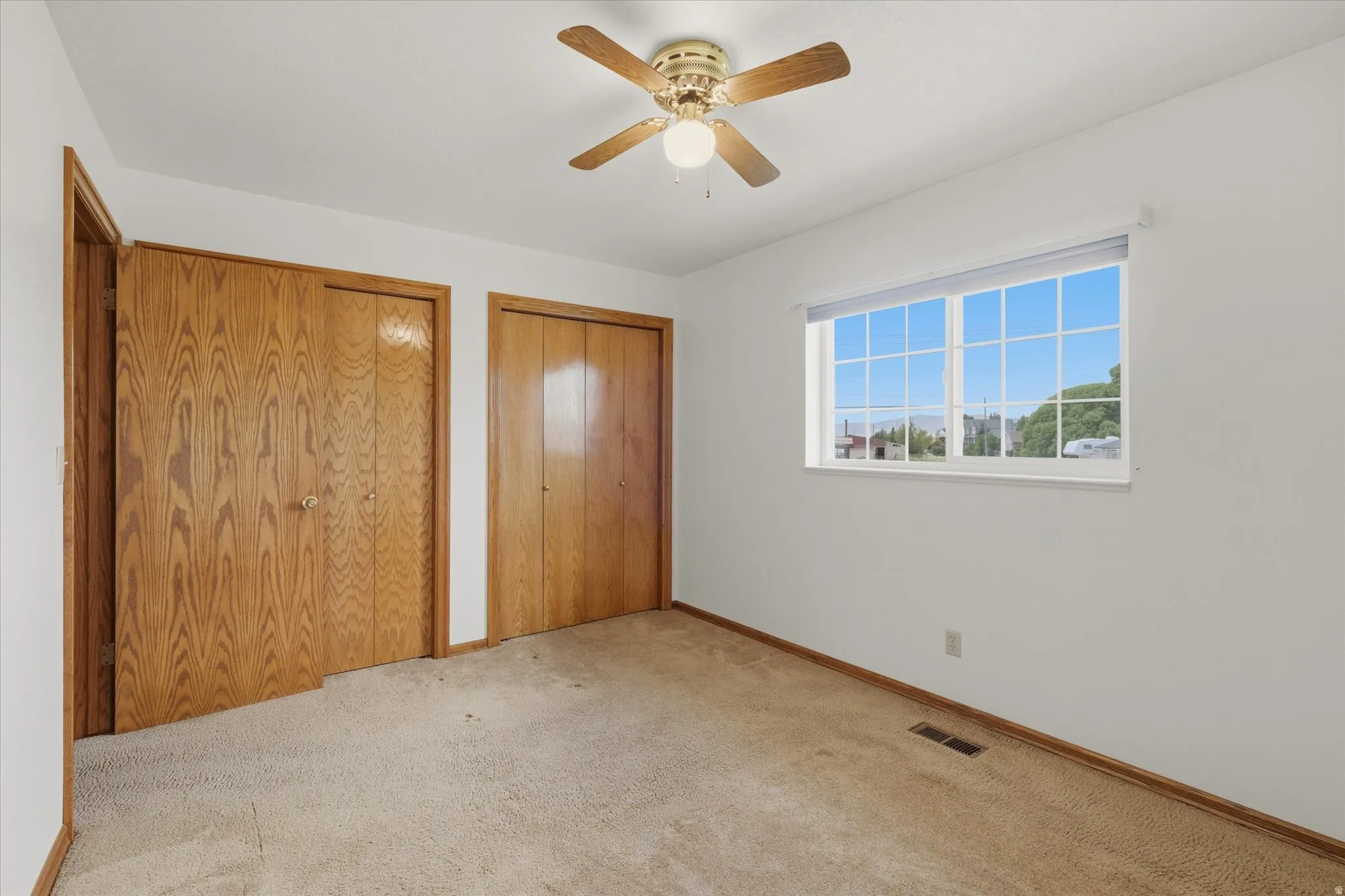 Carpeted interior space featuring a multi-pane window, ceiling fan with light fixture, and two wood-finish bi-fold closet doors