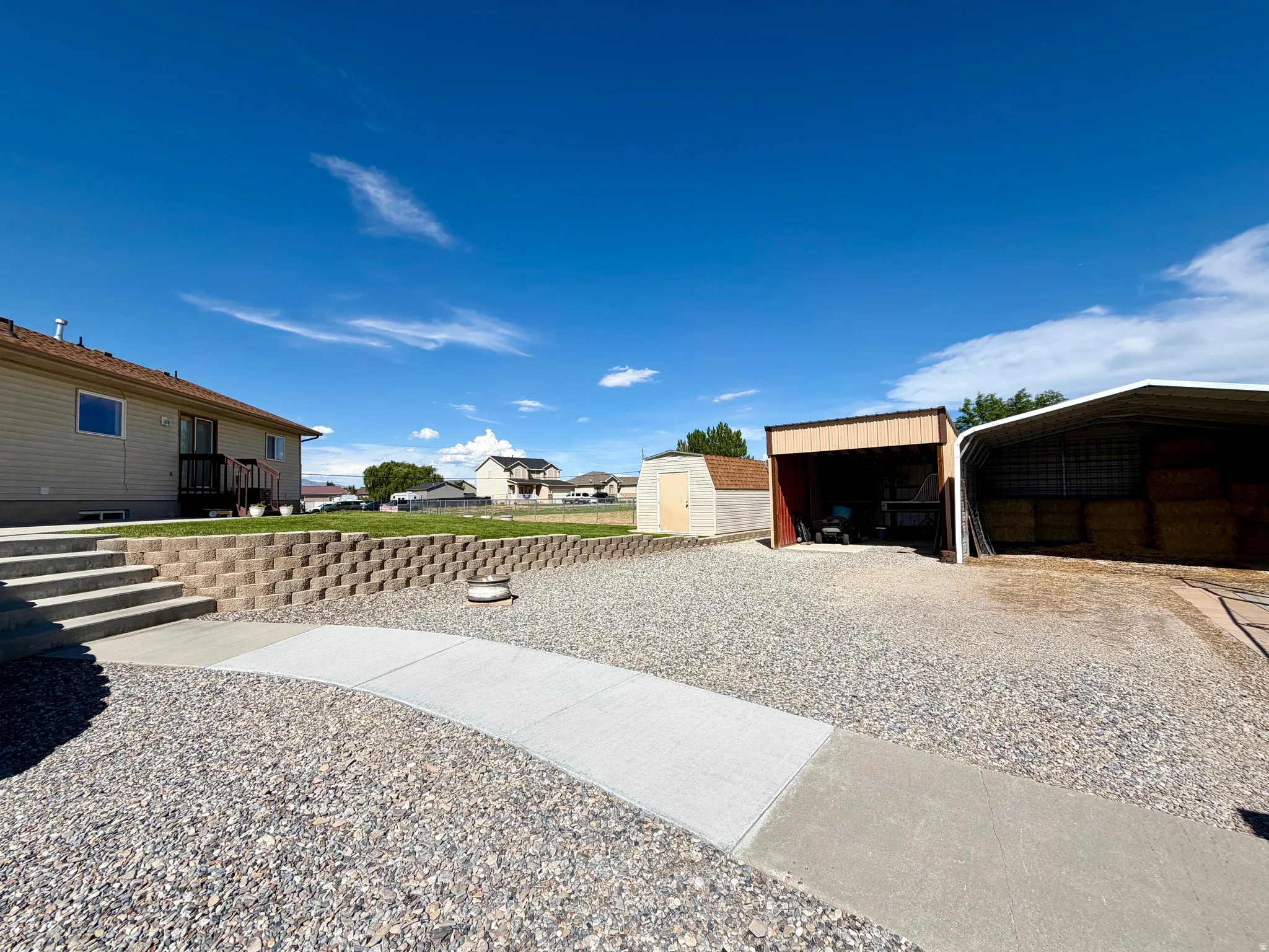 Expansive gravel yard with concrete walkways, a retaining wall, and multiple outbuildings, including a shed and two covered structures
