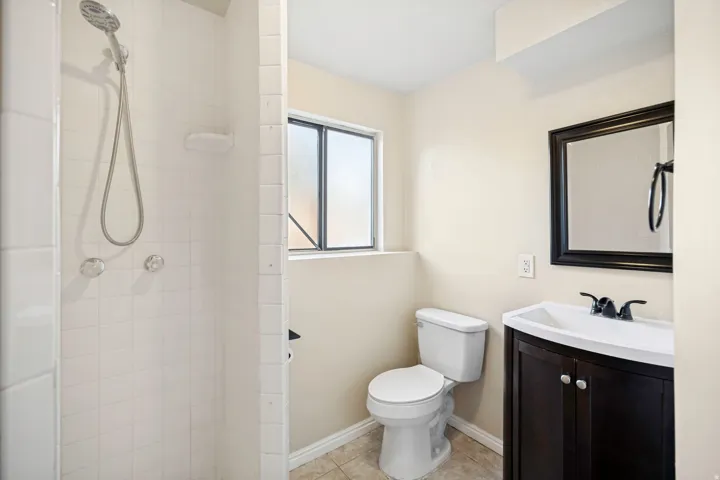Bathroom featuring a walk-in shower with white tile surround, a dark wood-finish vanity with an integrated sink, a dark framed mirror, and a window with obscured glass