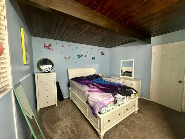 Room featuring a wood-panel ceiling with exposed beams, light blue painted walls, and textured carpet flooring