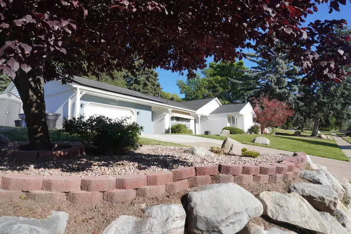 Landscaped front yard featuring a red brick retaining wall, natural rock formations, mature trees, and a concrete driveway