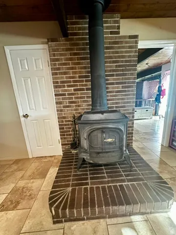 Cast iron wood-burning stove with a brick surround and hearth, featuring a dark metal stovepipe extending to a wood-beamed ceiling