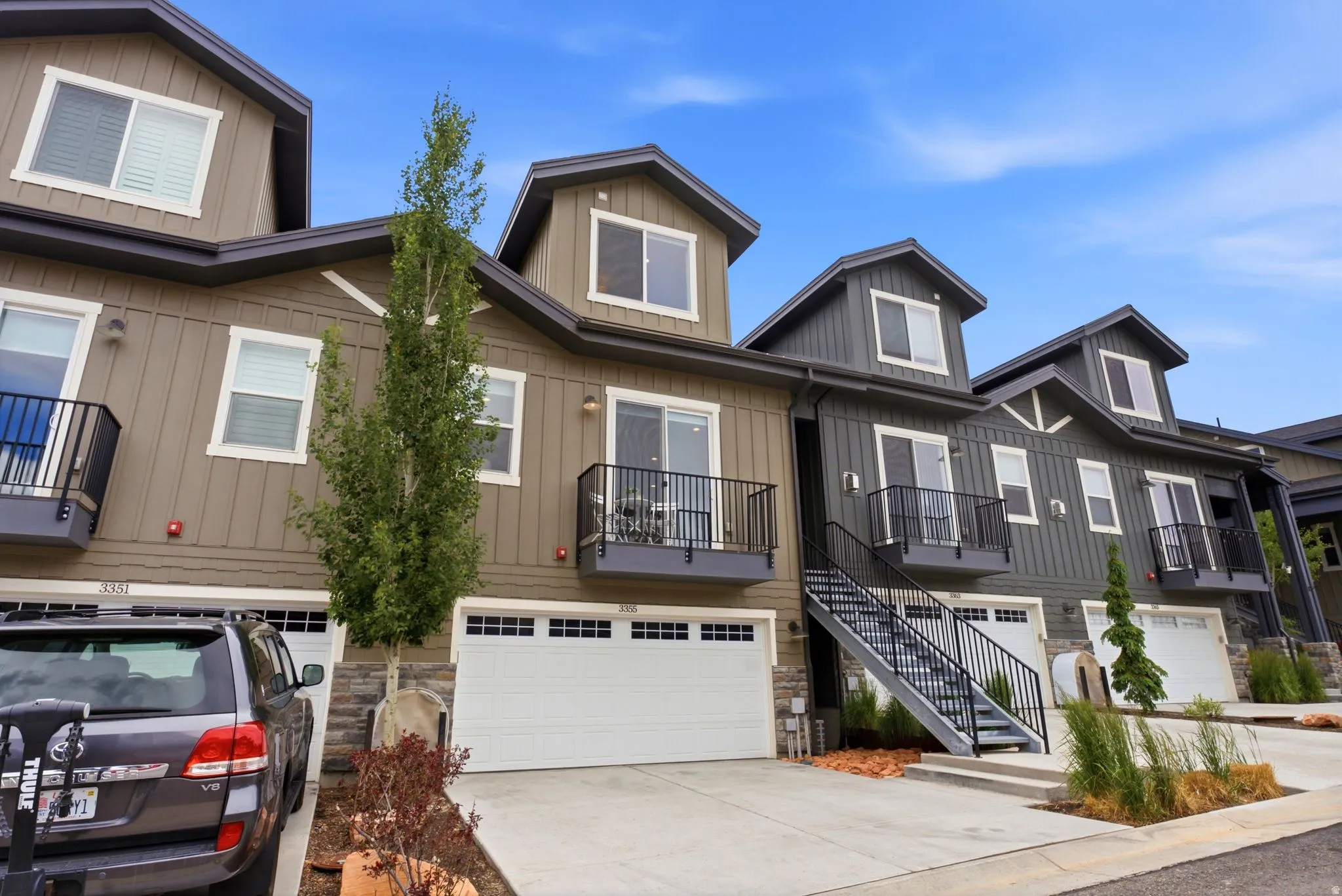 Modern townhome exterior featuring contemporary siding, individual garages, private balconies with black railings, dormer windows, and a dedicated exterior staircase