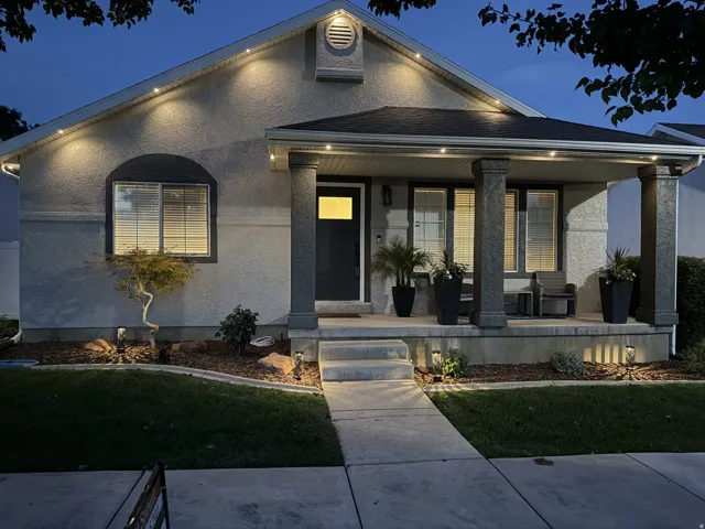 Stucco exterior with a covered porch featuring two columns, recessed lighting, concrete steps, and a landscaped front yard with ground lighting
