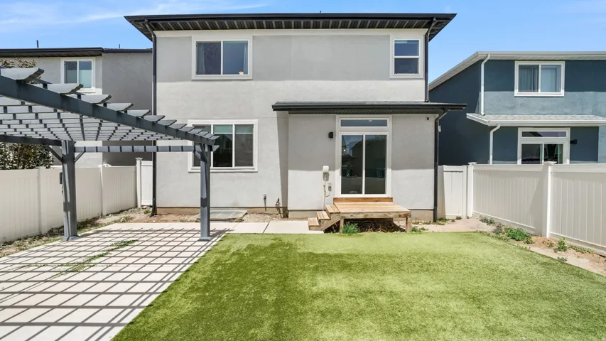 Rear exterior featuring a sliding glass door, an artificial turf lawn, a wooden deck, a pergola over a paved patio, and white privacy fencing